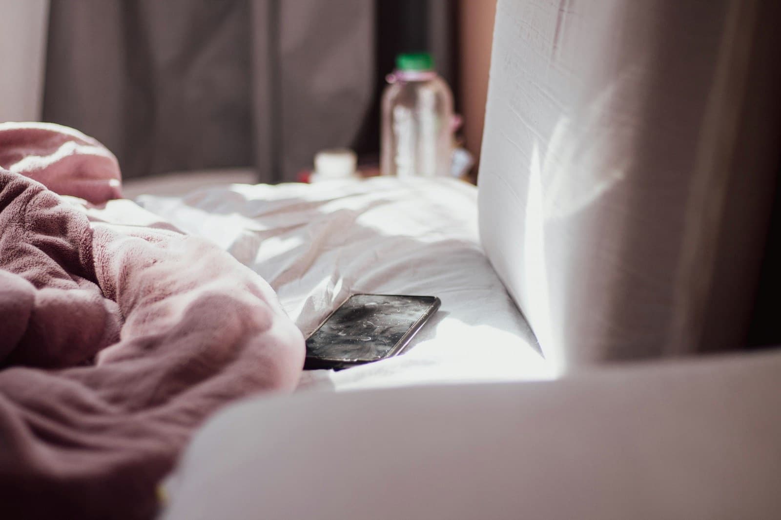 A phone resting on white bedsheets beside a pink blanket in morning sunlight