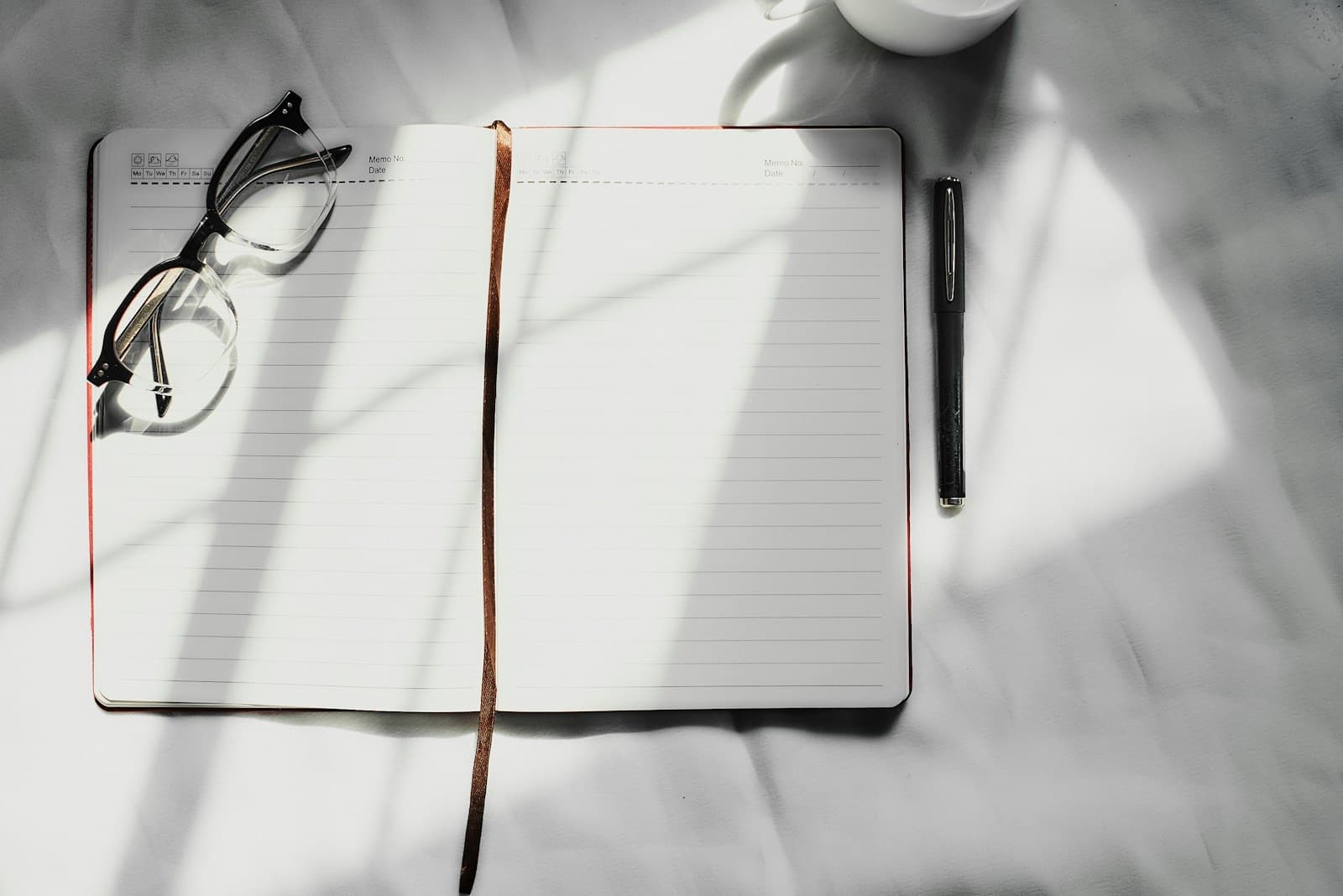 Open blank journal with glasses, a pen, and a cup of coffee in window light