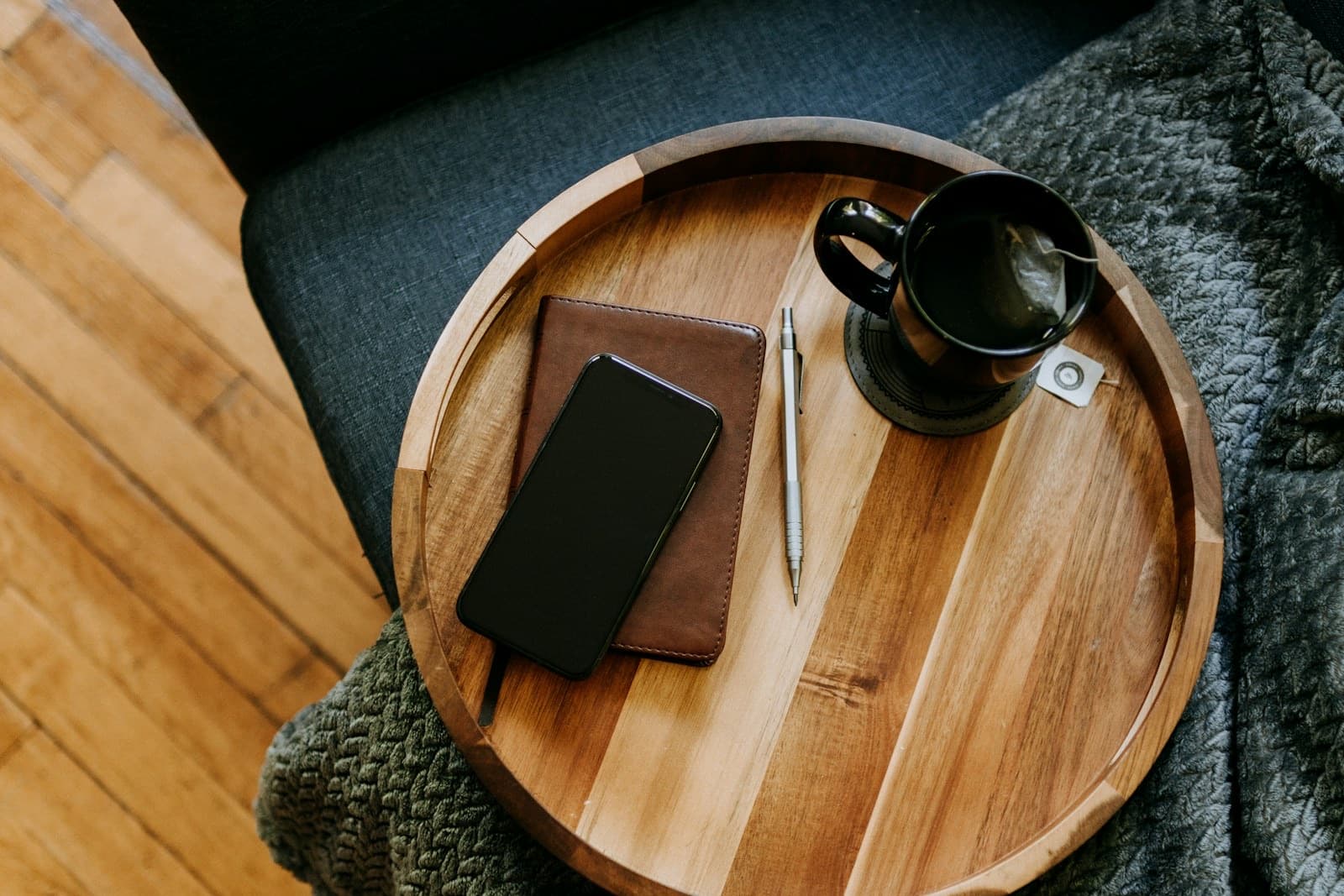 Top-down view of a wooden tray with a phone, a closed notebook, a pen, and a mug of tea