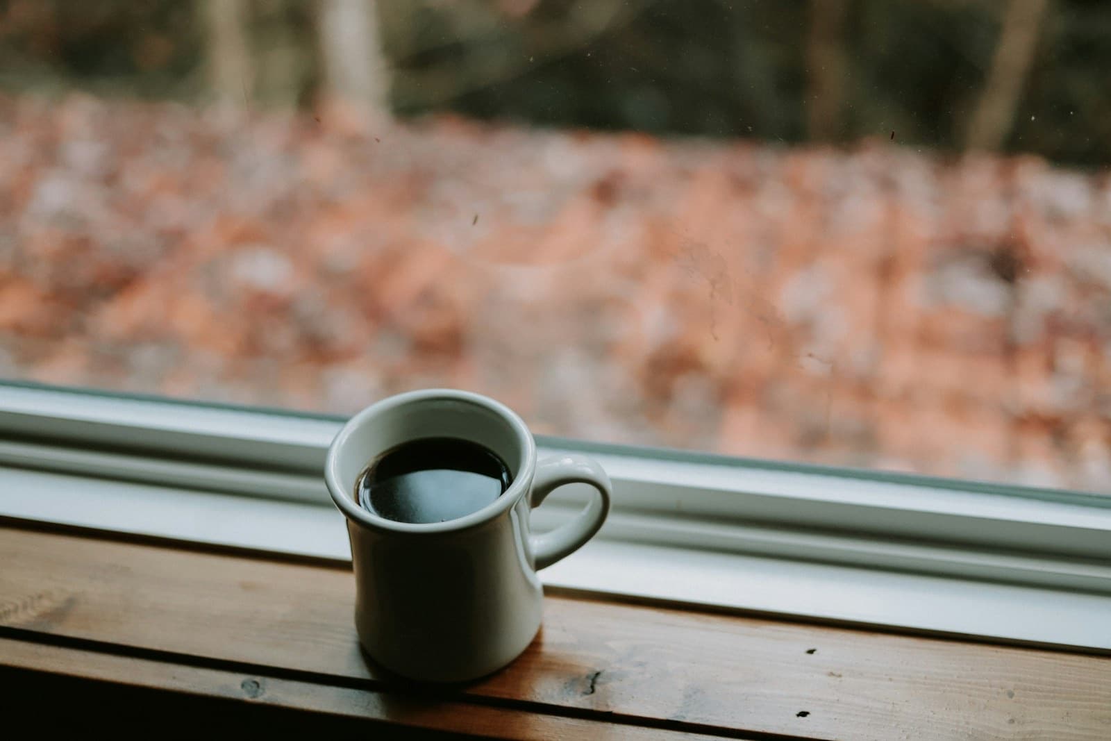 White mug of coffee on a wooden windowsill with autumn trees outside