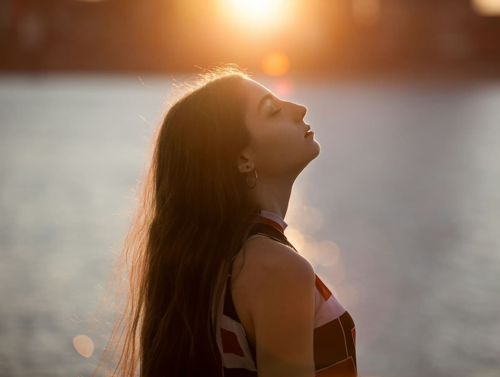 Woman with her eyes closed, face turned toward golden-hour light beside the water