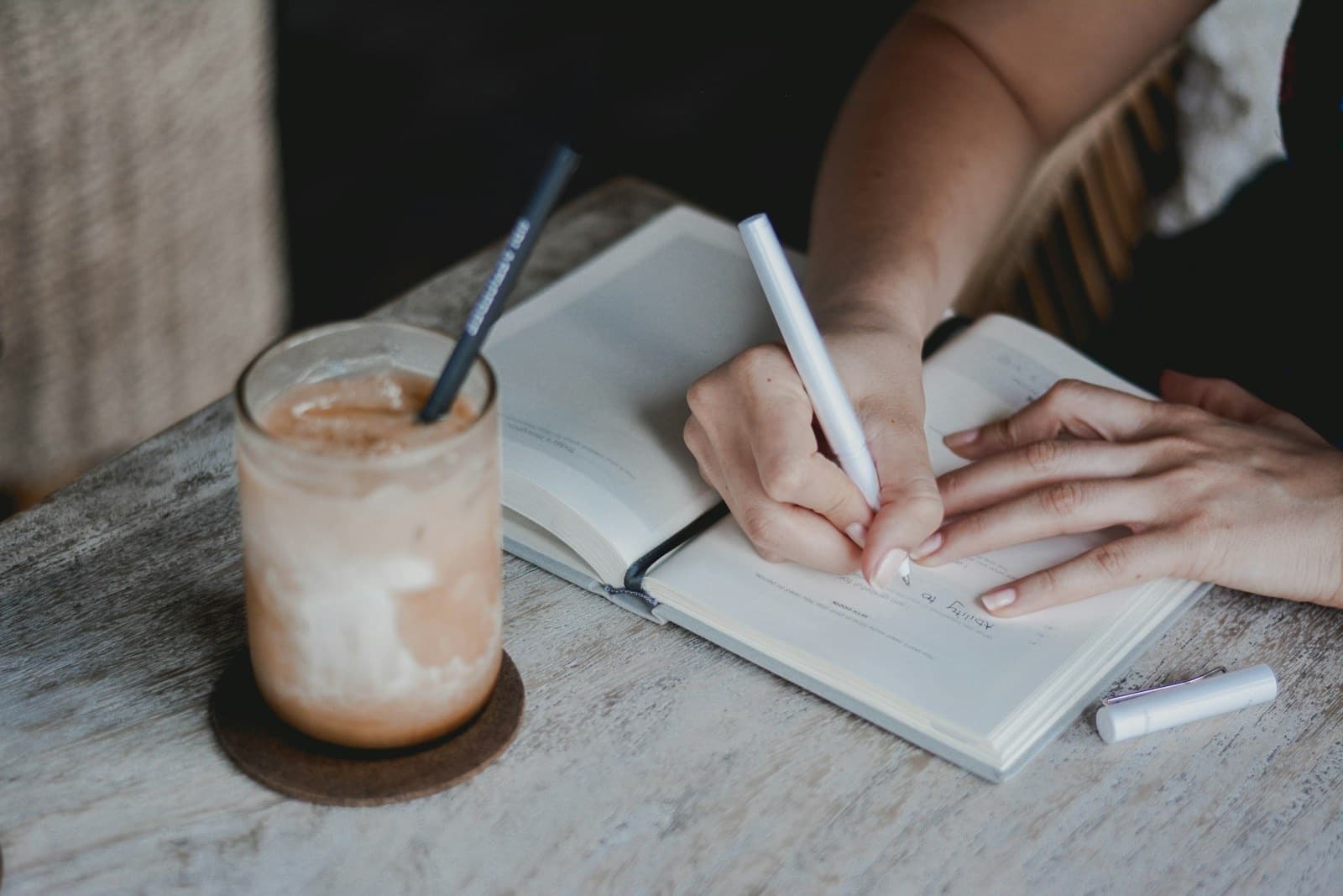 Close-up of a hand writing in a journal beside an iced coffee