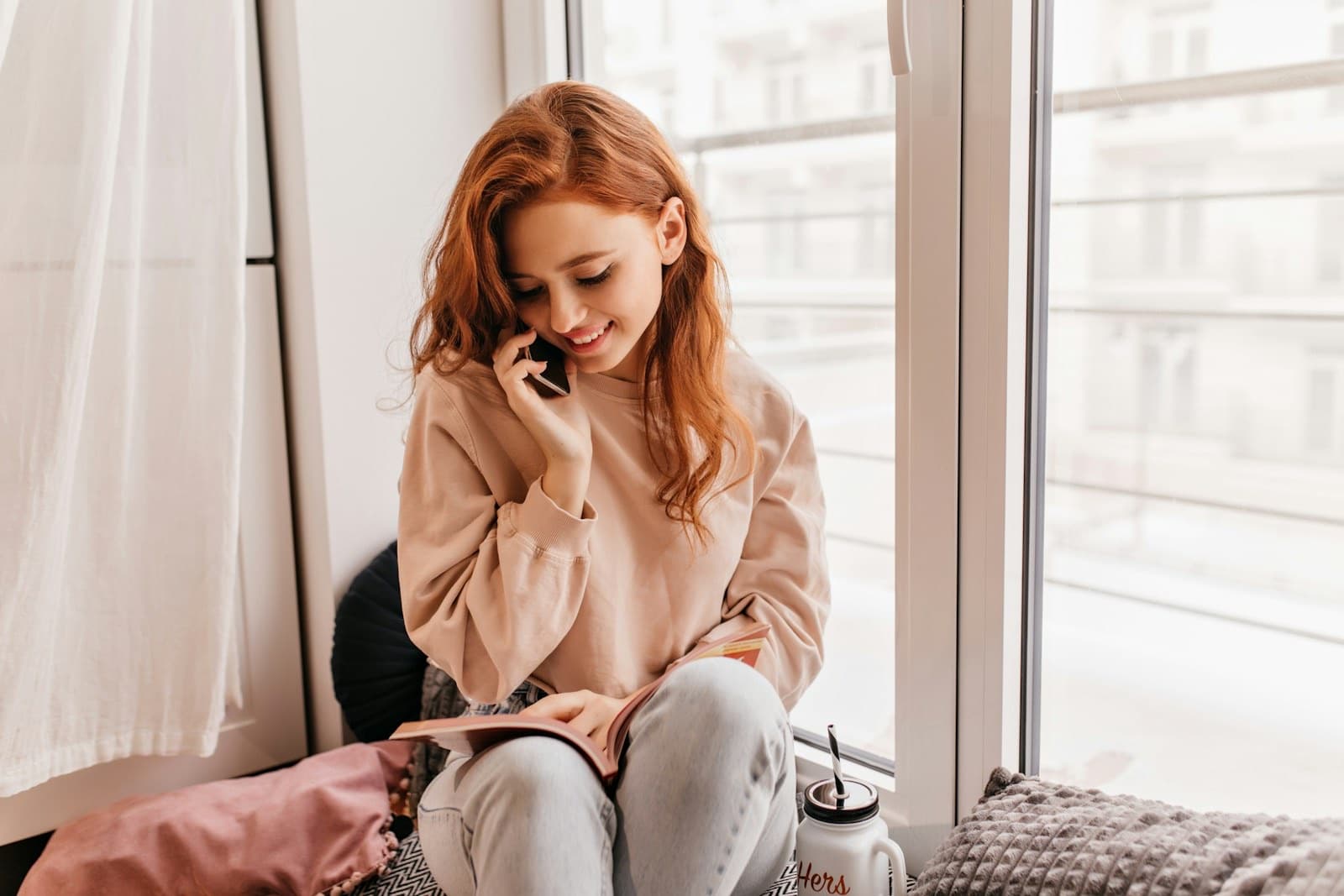 Woman in a beige sweater on a window seat, smiling softly during a phone call
