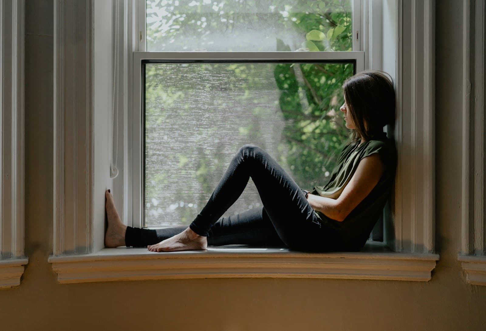 Woman sitting alone on a window sill in soft daylight, looking out
