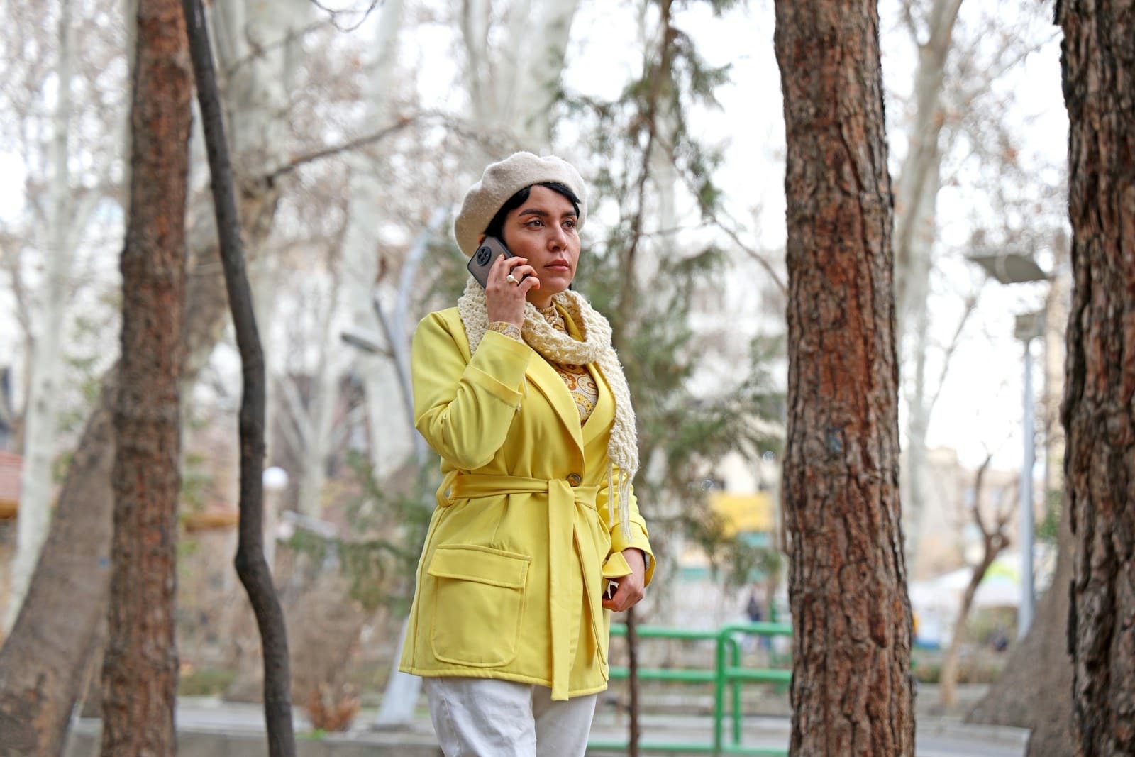 Woman in a yellow coat talking on her phone while walking through a winter park
